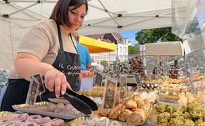 Verkaufsstand auf einem Markt: Eine Händlerin bereitet frisches Mandelgebäck zu, im Hintergrund sind weitere Marktstände und Besucher zu sehen