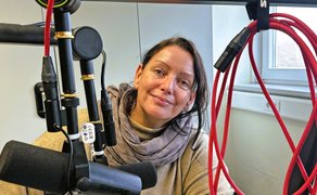 Frau mit braunem Schal sitzt vor einem schwarzen Mikrofon, im Hintergrund Fenster und rote Kabel an der Wand.
