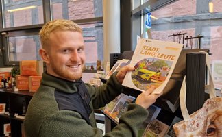 Person hält mehrere Ausgaben der Zeitschrift »Stadt, Land, Leute« mit Titelbild eines gelben Autos und Personen in Kostümen.