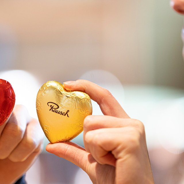 Zwei Rausch-Schokoladenherzen in Rot und Gold werden im Schokoladenhaus Peine in den Händen gehalten und symbolisieren feinen Schokoladengenuss aus der Chocolaterie Peine. Die Nahaufnahme zeigt hochwertige Süßwaren von Rausch als beliebtes Geschenk und Genussmoment in Peine.