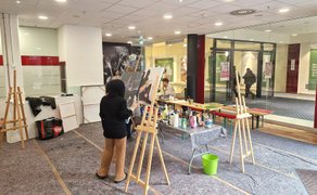 Ein Mädchen arbeitet an einem Bild auf einer Staffelei in einem Geschäftsraum. Am Schaufenster laufen Passanten vorbei. 