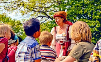Eine Gruppe von Kindern steht im Freien unter grünen Bäumen und hört aufmerksam einer Frau zu, die eine historische Stadtführung durchführt. Die Frau trägt ein mittelalterlich wirkendes Kleid mit Kopftuch und spricht lebendig zu den Kindern, die sich neugierig um sie versammelt haben. Die Szene wirkt freundlich und lebendig bei sonnigem Wetter.