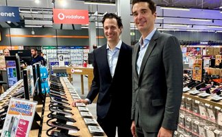 Zwei Männer stehen lächelnd nebeneinander in einem Elektronikmarkt. Im Vordergrund und Hintergrund sind verschiedene ausgestellte Elektrogeräte zu sehen.