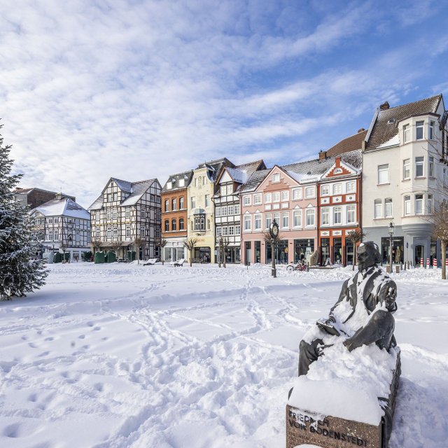 Verschneiter Marktplatz in Peine mit historischer Häuserzeile, Winterbaum und Statue im Vordergrund bei klarem Winterhimmel.