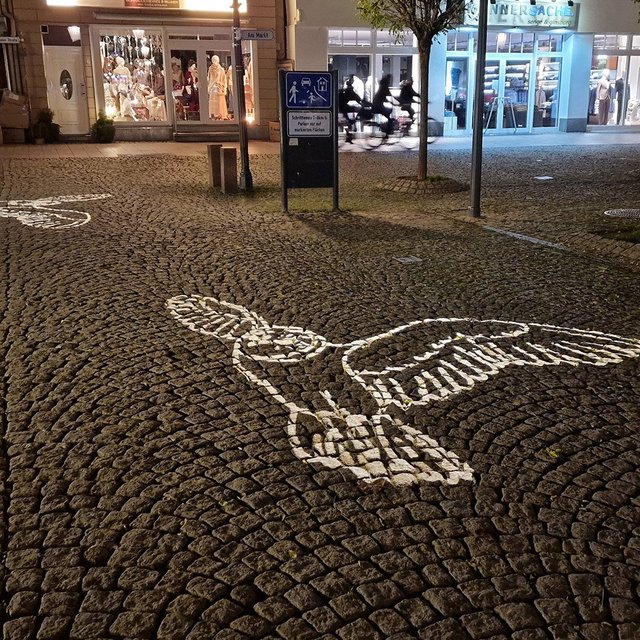 Was fliegt denn da über den Historischen Marktplatz in Peine? Leuchtende Eulen setzen seit einigen Tagen dort stimmungsvolle Akzente. Energiesparende LED-Projektoren lassen mit Einsetzen der Dunkelheit die Motive des Peiner Wahrzeichens auf dem Pflaster erscheinen. Die Installation findet großen Anklang: Immer wieder bleiben Besucher der Innenstadt stehen, um Fotos zu machen – und vor allem Kinder reagieren neugierig auf die leuchtenden Figuren. Für die neue Projektion wurden zwei Masten errichtet, die in Kürze zusätzlich mit Fahrradständern ergänzt werden – rechtzeitig zum Peiner Weihnachtsmarkt.  Weitere Projektoren schaffen mit stilisierten Baumkronen Atmosphäre an der St.-Jakobi-Kirche, deren Turm außerdem von neuen LED-Strahlern erhellt wird. Diese tragen erheblich zur Energieeinsparung bei. Zur bevorstehenden Weihnachtszeit können sich Besucher der Innenstadt außerdem auf eine Erweiterung der Beleuchtung in der Lindenstraße freuen.  Illumination schafft stimmungsvolle Erlebnisse Bürgermeister Klaus Saemann ist überzeugt von den positiven Auswirkungen der Lichtinszenierungen in der Innenstadt: „Wir sind sehr dankbar über die finanzielle Unterstützung des Bundes im Rahmen Förderprogramms ‚Zukunftsfähige Innenstädte und Zentren‘ (ZIZ). Ohne die Förderung hätten wir angesichts der schwierigen Finanzlage der Stadt das Maßnahmenpaket im Projekt PEINE2030 nicht umsetzen können. Gerade die Maßnahmen zur Illuminierung der Innenstadt sorgen für eine positive Stimmung beim Aufenthalt in der Fußgängerzone in der dunkleren Jahreszeit sowie das ganze Jahr hinweg in den Nacht- und Abendstunden. Ich freue mich, dass dies Menschen aus den Nachbarkommunen und der Region Anlass zu einem Besuch der Peiner Innenstadt gibt.“  Die ZIZ-Projekte wurden in enger Zusammenarbeit zwischen der Stadtverwaltung und der Peine Marketing umgesetzt. Anja Barlen-Herbig, Geschäftsführerin der Peine Marketing ist ebenfalls begeistert von der neuen Illuminierung: „Das ist ein weiterer wichtiger Baustein, der zur Attraktivierung der Innenstadt in den Abendstunden beiträgt. Die Illuminierung war ein Wunsch vieler Innenstadtanrainer und ein Projektergebnis des Beteiligungsprozesses ‚Zukunftswerkstatt‘, den wir durchgeführt haben. Die neue Illuminierung zahlt auf die Identität der Stadt ein und schafft in den Abendstunden ein wunderbares Erlebnis.“ Was ist das Ziel des Förderprogramms ZIZ? Im Zusammenhang mit dem Förderprogramm Zukunftsfähige Innenstädte und Zentren (ZIZ) setzt die Stadt Peine seit 2023 Maßnahmen zur Attraktivitätssteigerung der Innenstadt unter dem Titel PEINE2030 um. Dazu gehören der Ausbau der Digitalisierung und des Citymanagements, der umfangreiche Markenprozess, Projekte für jüngere Zielgruppen wie das innovative Sitzmobiliar an der Töpfers Mühle, die prachtvollen Blumenkübel oder die sogenannte Fotopoints – beleuchtete Figuren, die bei der Weihnachtsstadt Peine eine besondere Anziehungskraft auf die Besucher der Innenstadt haben. Die Illumination hat im ZIZ-Projekt von Beginn an eine große Rolle gespielt. Neben Sicherheitsaspekten dient die Illumination von Innenstädten der Schaffung einer positiven Atmosphäre und der Steigerung der Aufenthaltsqualität. Mit Hilfe der Fördergelder wurde bereits in der Weihnachtszeit der Jahre 2023 und 2024 die bestehende Weihnachtsbeleuchtung erweitert, darunter LED-Kugeln in den Baumkronen.