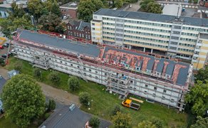Luftaufnahme eines langgestreckten Wohngebäudes mit Gerüst und Solarpaneelen auf dem Dach, umgeben von Bäumen und weiteren Gebäuden.