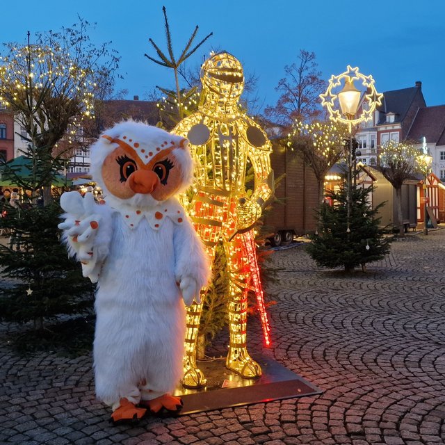 Uhlinchen steht auf dem weihnachtlich beleuchteten Marktplatz in Peine neben einer leuchtenden Ritterfigur, umgeben von Tannenbäumen und Weihnachtsbeleuchtung.