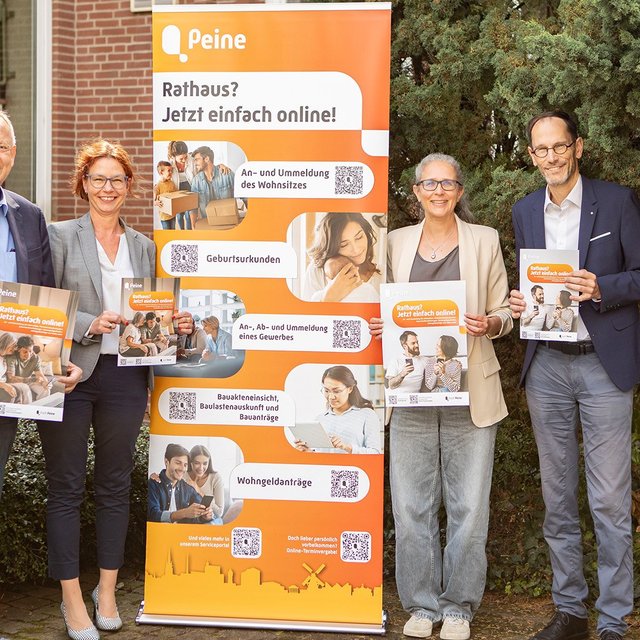 Vier Personen stehen vor einem Gebäude neben einem orangefarbenen Aufsteller zum digitalen Rathaus der Stadt Peine und halten Informationsmaterial in den Händen.