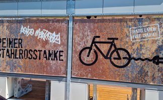 Rostfarbene Metalltafeln an einer Fahrradstation mit der Aufschrift „Peiner Stahlrosstanke“, einem Fahrradsymbol mit Stecker und dem Hinweis „Gratis laden / Free charge“.