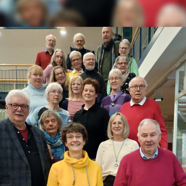 Gruppe von Menschen unterschiedlichen Alters in einem Innenraum mit Treppe und Geländer im Hintergrund.