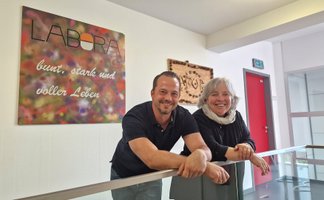 Ein Mann und eine Frau lehnen an einem Geländer, dahinter eine bunte Wandtafel mit der Aufschrift „LABORA bunt, stark und voller Leben“.