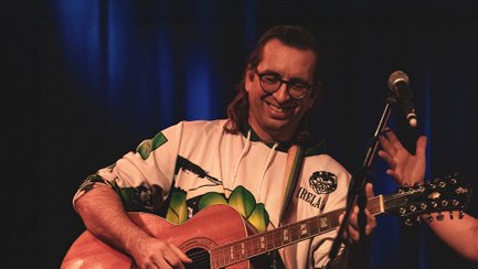 Musiker Fredy Hollschuh steht mit einer Akustikgitarre auf einer Bühne und lächelt während seines Auftritts. Neben ihm befindet sich ein Mikrofon, im Hintergrund sorgen dunkle Vorhänge und blaues Bühnenlicht für Konzertatmosphäre.