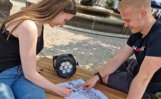 Zwei junge Erwachsene sitzen an einem Tisch im Freien, betrachten einen Lageplan und besprechen gemeinsam die Platzierung eines technischen Scheinwerfers.