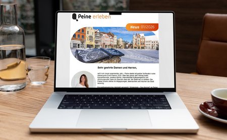 Laptop auf Holztisch zeigt Newsletter mit Überschrift „Peine erleben“ und einem Foto eines historischen Marktplatzes.