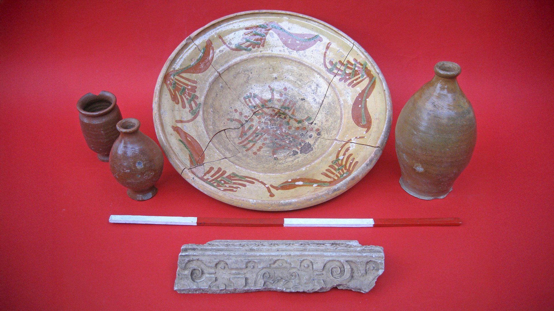 Vier archäologische Keramikgefäße und ein verziertes Steinfragment auf rotem Untergrund.