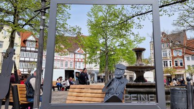 Sitzbank mit Metallfigur und Bilderrahmen-Installation auf dem Peiner Marktplatz mit Blick auf Brunnen und historische Fassaden als Stadtmöblierung im urbanen Stadtraum von Peine, Niedersachsen