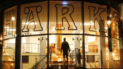 Blick durch die Glasfassade der Härke Braumanufaktur Peine auf die beleuchteten Kupferkessel im Inneren der Brauerei. Eine Person betritt den zentralen Brauraum und unterstreicht die moderne Verbindung von Braukunst, Architektur und Biergenuss aus Peine.