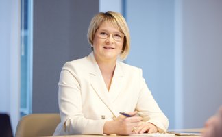 Frau mit kurzem blonden Haar in weißem Blazer sitzt an einem Tisch und hält einen Stift in der Hand.