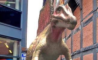 Augmented-Reality-Darstellung eines Dinosauriers in einer städtischen Umgebung zwischen modernen und historischen Gebäuden.