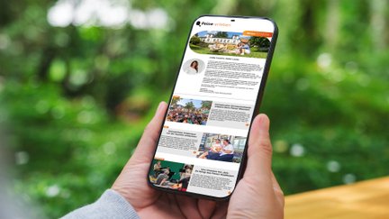 Eine Person hält ein Smartphone in der Hand. Auf dem Display ist der digitale Newsletter „Peine erleben“ geöffnet, der mit Bildern und Beiträgen zu Veranstaltungen, Freizeitangeboten und Erlebnissen in Peine gefüllt ist. Im Hintergrund ist eine unscharfe grüne Parklandschaft zu sehen.
