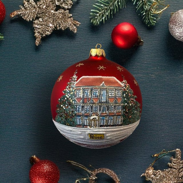 Rote Weihnachtskugel mit bemaltem Haus und Tannenbäumen, umgeben von Tannenzweigen und weiteren roten und silbernen Kugeln.