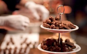 Zweistöckige Etagere mit verschiedenen Pralinen, im Hintergrund Hände mit weißen Handschuhen beim Schokoladenherstellen.