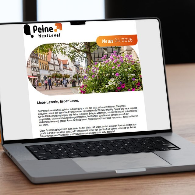 Laptop auf einem Schreibtisch mit geöffneter digitalen Ausgabe des Newsletters „Peine NextLevel News 04/2026“. Auf dem Bildschirm ist ein Beitrag zur Entwicklung der Peiner Innenstadt mit einem Foto der Innenstadt und blühenden Blumen zu sehen. Neben dem Laptop stehen ein Coffee-to-go-Becher, ein orangefarbenes Notizbuch und ein Schreibblock.