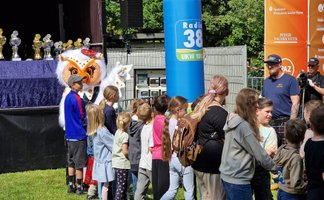 Uhlinchen steht beim Highland Gathering in Peine zwischen vielen Kindern vor der Bühne im Stadtpark. Im Hintergrund sind Pokale, schottische Dekorationen und Veranstaltungsbanner zu sehen. Die Kinder warten gespannt auf das gemeinsame Tauziehen bei den Kinder-Games.