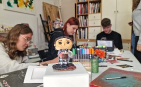 Eine kleine Skulptur im Vordergrund, im Hintergrund arbeiten Jugendliche an einem Tisch mit Malutensilien.