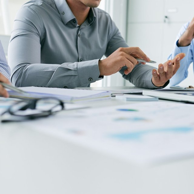 Mehrere Personen sitzen an einem Tisch in einer Besprechung, diskutieren Unterlagen und arbeiten gemeinsam mit Laptop und Notizen in einer professionellen Büroumgebung.