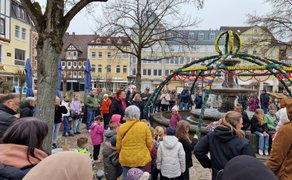 Eine größere Gruppe von Menschen, darunter viele Kinder und Erwachsene, versammelt sich auf einem Marktplatz rund um einen geschmückten Brunnen. Der Brunnen ist mit grünen Girlanden und bunten Ostereiern dekoriert. Einige Kinder stehen vorne und schauen aufmerksam zu, während Erwachsene im Hintergrund beobachten. Im rechten Bildbereich ist eine als Osterhase verkleidete Person zu sehen. Um den Platz herum stehen Bäume und historische Gebäude.