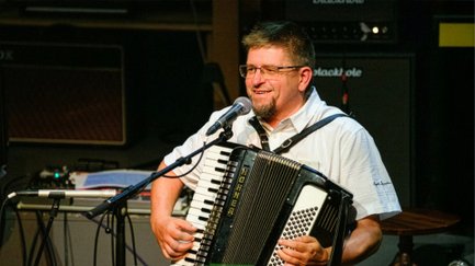 Musiker Rüdiger Schiller steht mit einem Akkordeon auf einer Bühne und singt lächelnd in ein Mikrofon. Im Hintergrund sind Lautsprecher und Bühnentechnik zu sehen, während die Szene in warmem Licht beleuchtet ist.