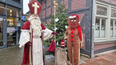 Zwei Figuren in Weihnachtskostümen stehen nebeneinander: eine als Bischof mit rotem Gewand und Mitra, die andere als Lebkuchenmann mit roter Mütze und roten Knöpfen.