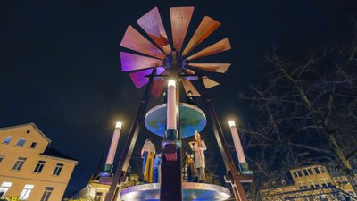 Bunte Weihnachts-Pyramide mit Figuren und beleuchteten Kerzen, umgeben von Gebäuden und dunklem Nachthimmel.