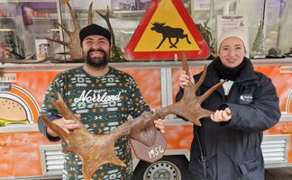 Ein Mann und eine Frau halten gemeinsam ein großes Geweih mit der Jahreszahl „1956“, im Hintergrund ein Warnschild mit Elch-Symbol an einem Foodtruck.