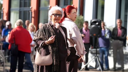 Zwei Models präsentieren Mode bei einer Modenschau im Freien auf dem Historischen Marktplatz in Peine. Im Vordergrund trägt eine ältere Frau ein elegantes dunkles Outfit mit gemustertem Schal und Handtasche, daneben steht ein weiteres Model mit roten Haaren in heller Jacke. Im Hintergrund verfolgen Besucher das Event in der Innenstadt.