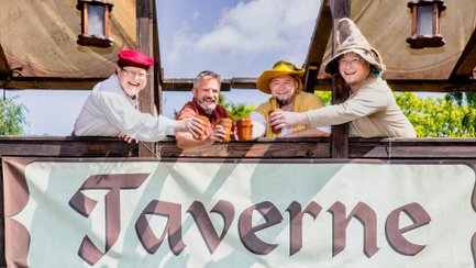 Vier Mitglieder der Mittelalter-Folk-Band Bierschmeichler stehen in historisch anmutender Gewandung auf einem hölzernen Balkon und stoßen lachend mit Tonbechern an. Unter ihnen hängt ein großes Banner mit mittelalterlicher Schrift, während über ihnen ein blauer Himmel mit Wolken zu sehen ist.