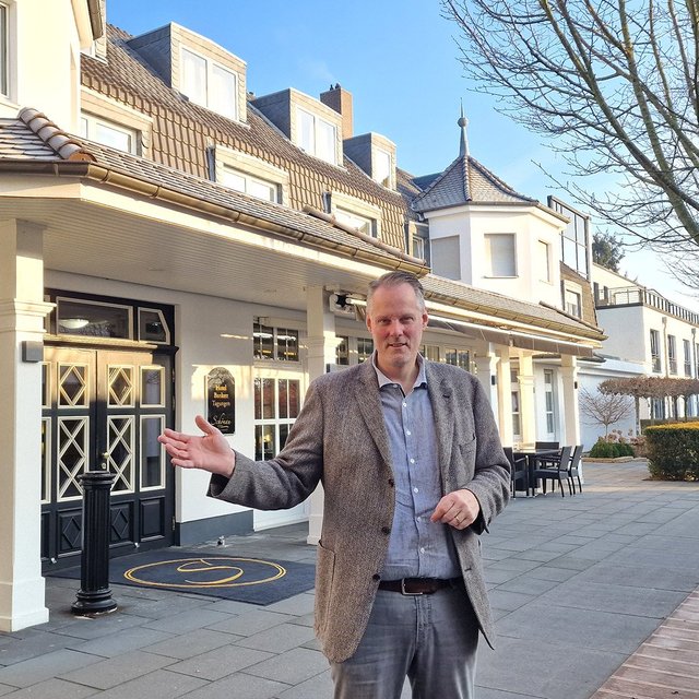 Mann in Sakko steht vor einem mehrstöckigen Hotelgebäude mit Innenhof.