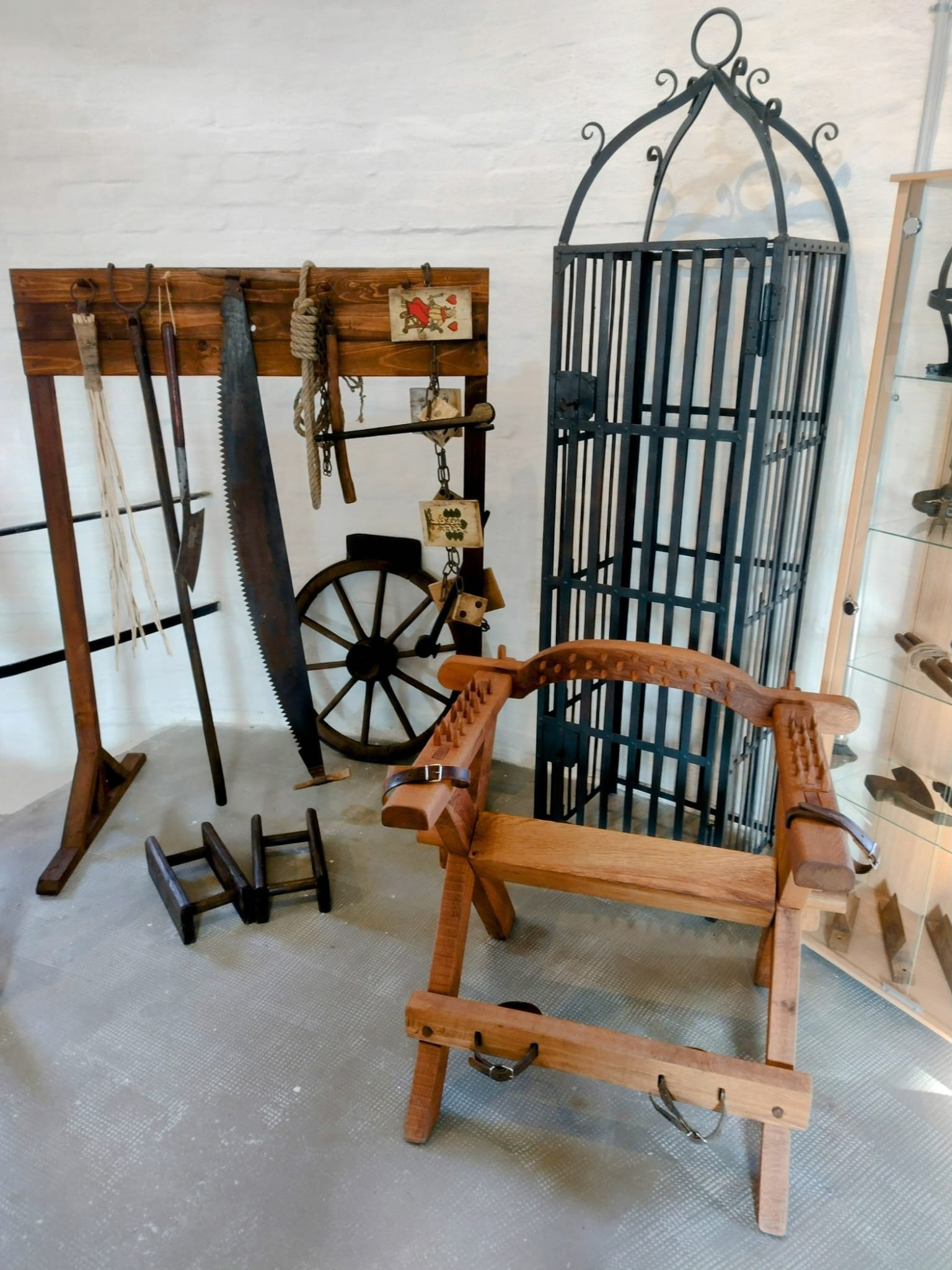 Ausstellung historischer Folterinstrumente mit Holzgestell, Säge, Seilen, Streckbank und Eisenkäfig in einem Museumsraum, die die Methoden der Justiz zur Zeit der Hexenverfolgung veranschaulichen.