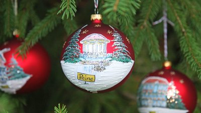 Rote Christbaumkugel mit winterlicher Landschaft und Schriftzug 'Peine' hängt an Tannenzweig neben einer einfarbigen roten Kugel.