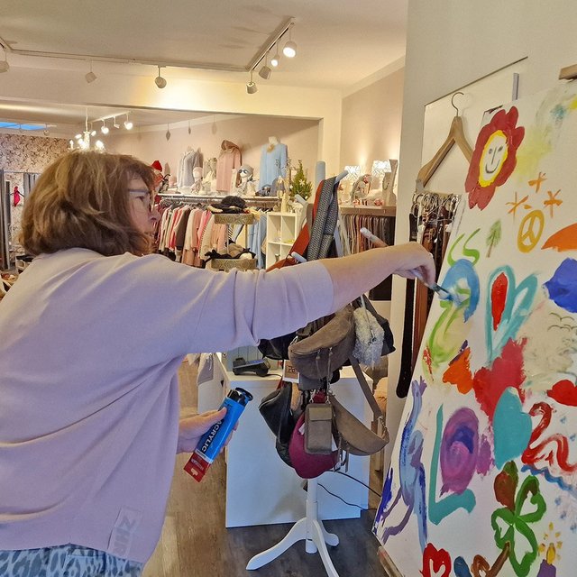 Frau malt mit Pinsel auf eine große Leinwand mit bunten, kindlich gemalten Motiven in einem Raum mit mehreren Personen und Kleidung an Ständern.