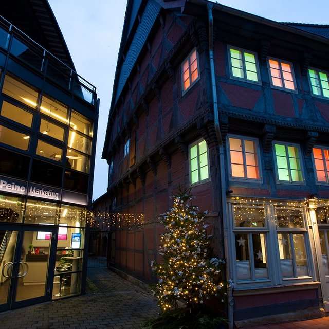 Beleuchtetes Fachwerkhaus und modernes Gebäude mit Glasfassade bei Dämmerung, geschmückt mit Weihnachtsbeleuchtung und Baum mit Lichterkette.