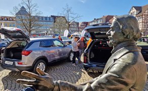 Bronzestatue mit ausgestrecktem Arm im Vordergrund vor mehreren geparkten Autos mit geöffneten Kofferräumen auf einem gepflasterten Marktplatz mit Fachwerkhäusern im Hintergrund