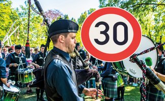 Ein Dudelsackspieler in traditioneller schottischer Kleidung spielt bei einem Highland Gathering, umgeben von einer Trommlergruppe und Zuschauern im Freien; im Vordergrund ist ein rundes Tempo-30-Verkehrsschild zu sehen.