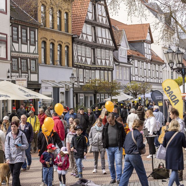 Viele Menschen besuchen eine belebte Fußgängerzone mit Fachwerkhäusern, Aktionsständen und orangefarbenen Luftballons in der Innenstadt von Peine.