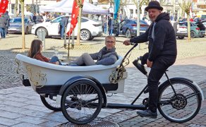 Dreirädriges Fahrrad mit einer Badewanne als Sitzgelegenheit, in der zwei Personen sitzen, eine dritte Person fährt das Fahrrad auf gepflastertem Platz