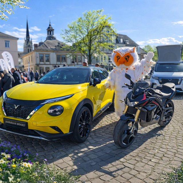Gelbes Auto und schwarzes Motorrad auf gepflastertem Platz mit Person im Eulen-Kostüm und mehreren Besuchern bei sonnigem Wetter.