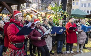 Gruppe von Menschen mit Weihnachtsmützen und Winterkleidung singt gemeinsam auf einem Weihnachtsmarkt vor festlich geschmückten Ständen.