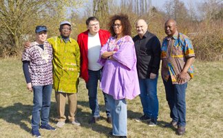 Die Band Pretty Cashanga steht mit sieben Personen auf einer Wiese und blickt in die Kamera. Einige tragen farbenfrohe Kleidung.