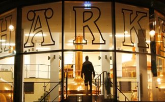 Blick durch die Glasfassade der Härke Braumanufaktur Peine auf die beleuchteten Kupferkessel im Inneren der Brauerei. Eine Person betritt den zentralen Brauraum und unterstreicht die moderne Verbindung von Braukunst, Architektur und Biergenuss aus Peine.